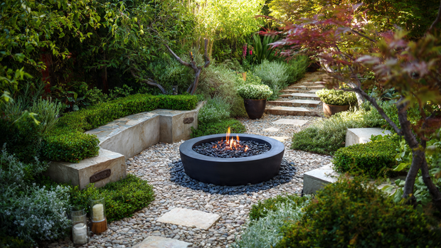 16 Cozy and Modern Backyard Fire Pit Ideas 13 Landscaped fire pit surrounded by plants, gravel, and decorative planters.
